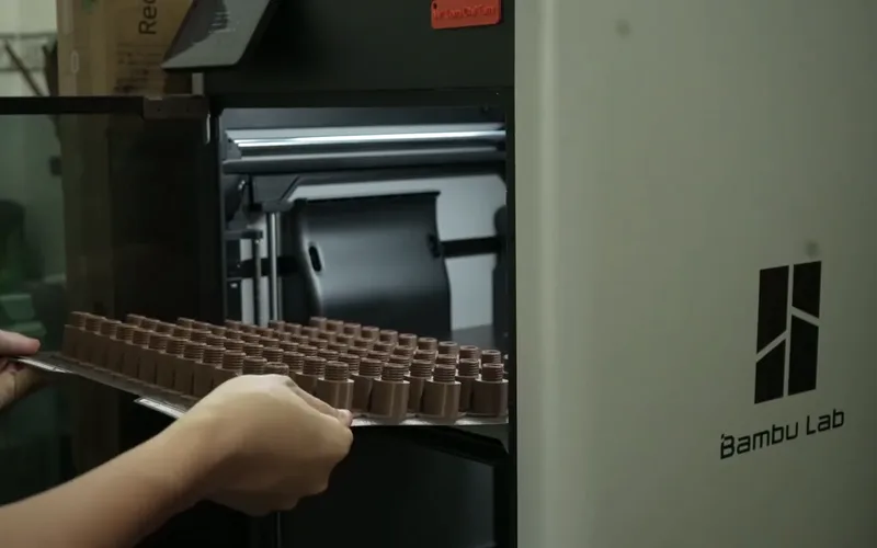 A hand lifting a print bed full of small brown parts out of a Bambu Lab printer